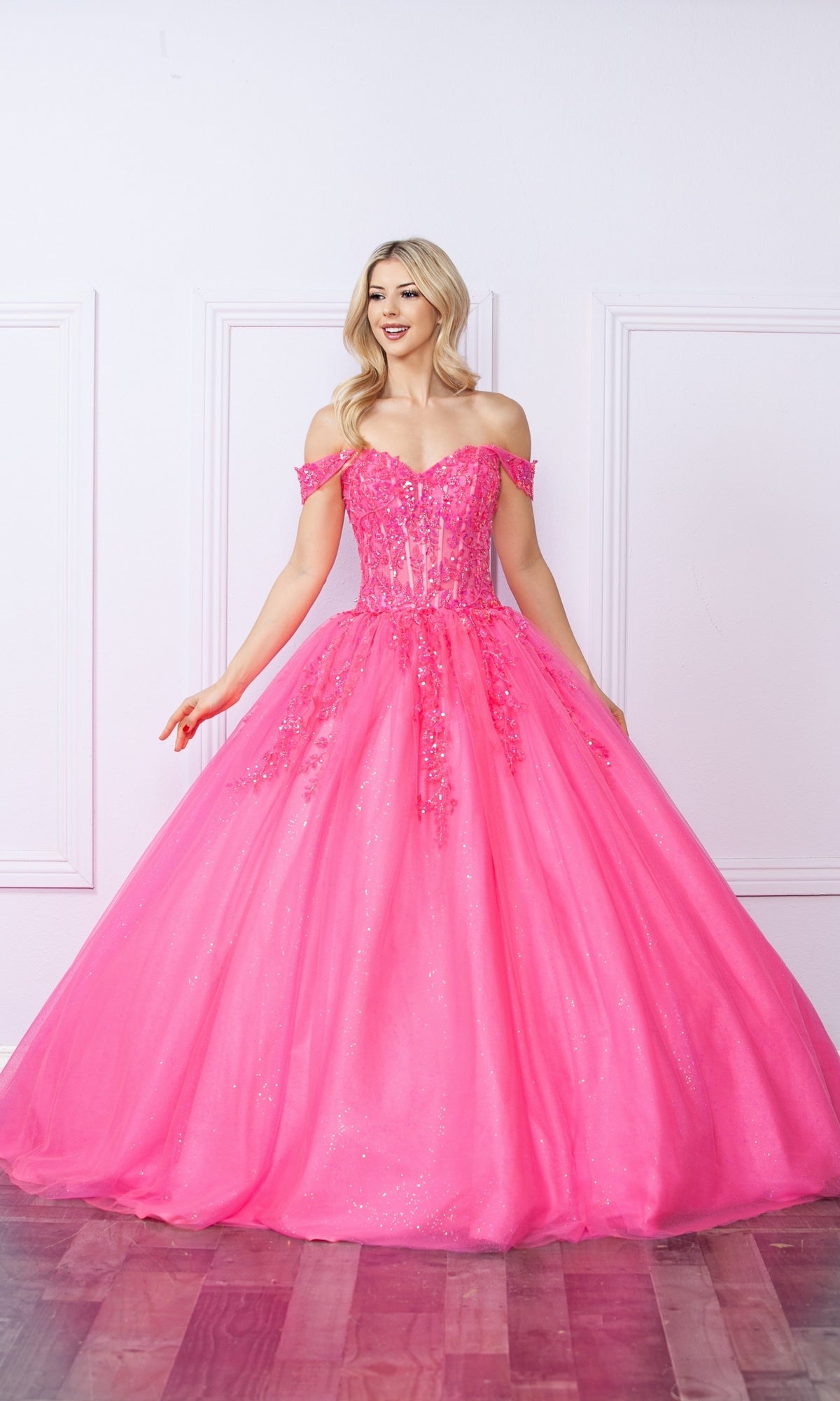 A woman stands indoors wearing the Bright Off-the-Shoulder Long Prom Ball Gown H1349, a vibrant pink dress with a full tulle skirt and sparkling sequins—an eye-catching choice for any formal event.